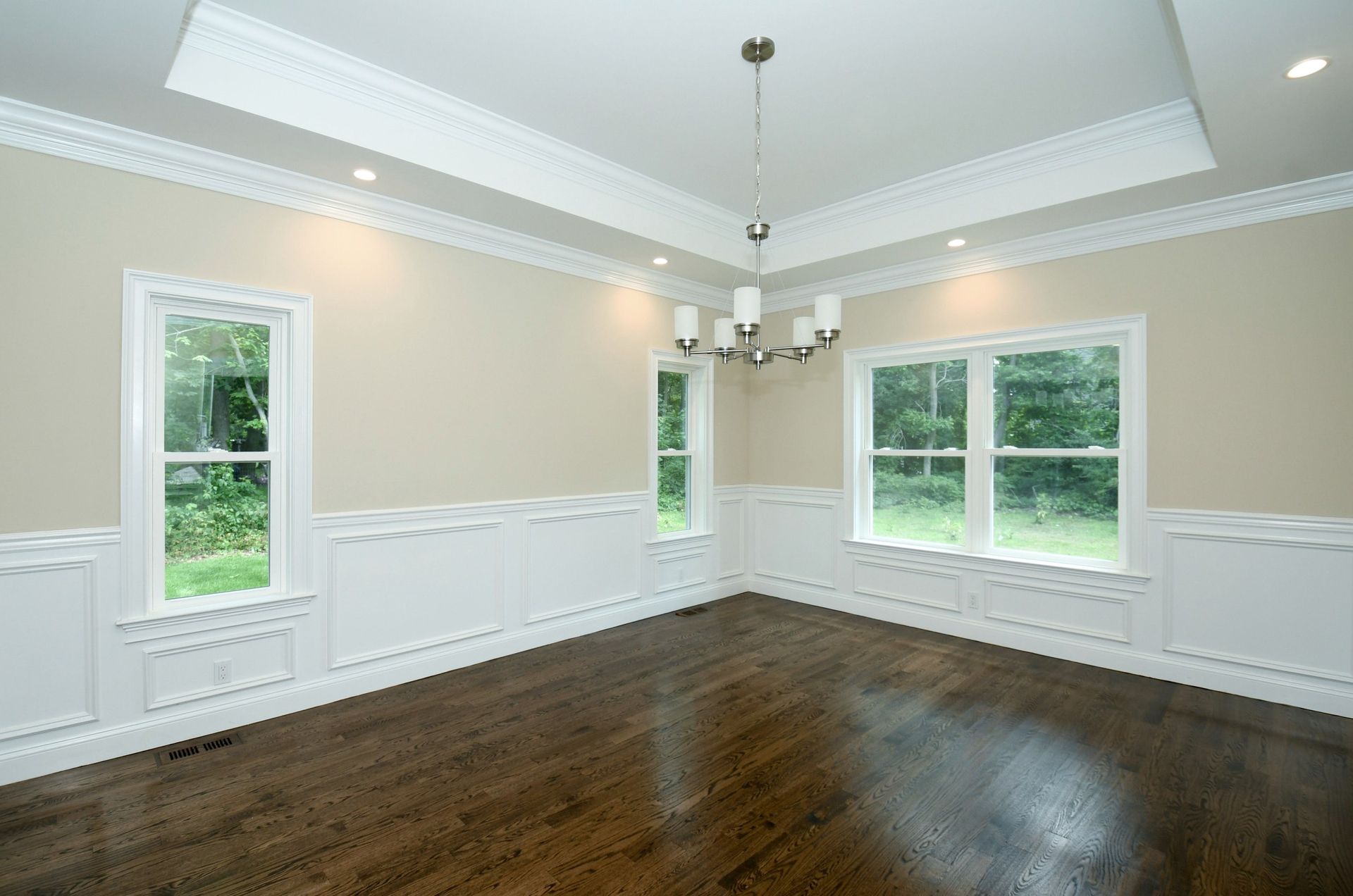 Empty dining room with dark hardwood floors, light tan walls, and white trim. A chandelier hangs from the recessed ceiling.