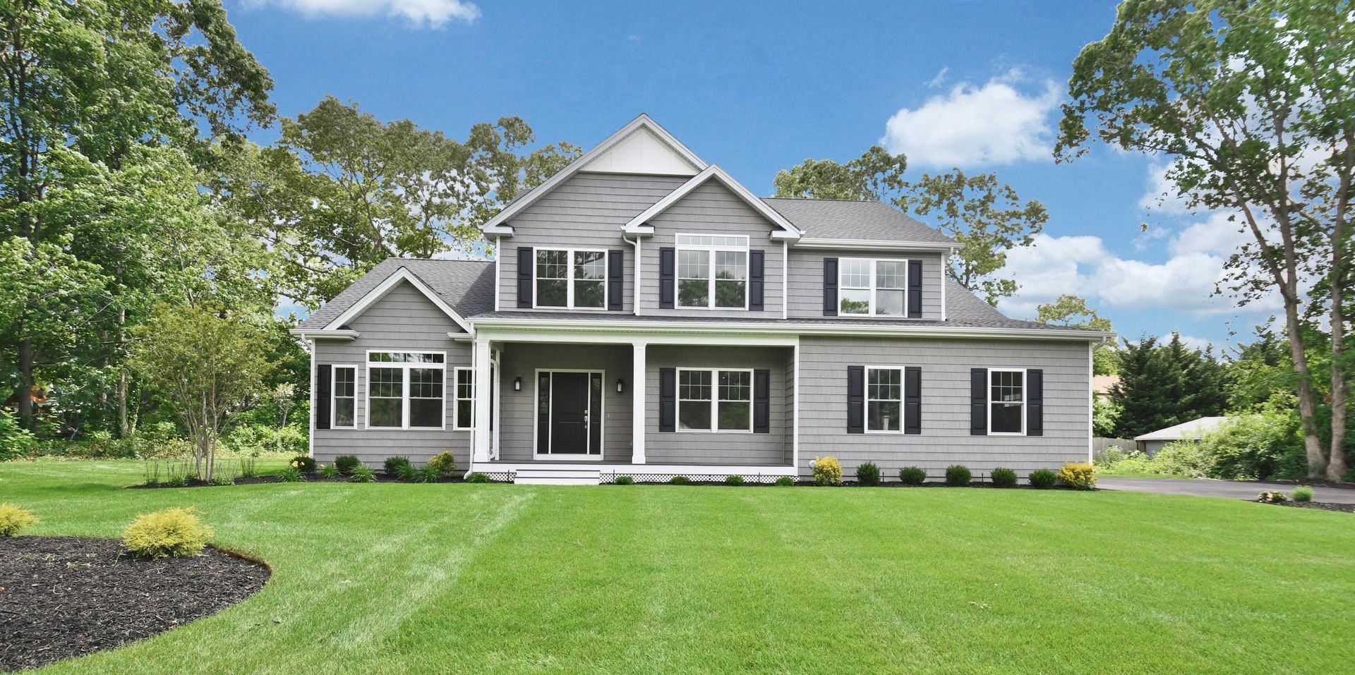 Two-story gray house with black shutters, a green lawn, and a blue sky. Trees surround the house.