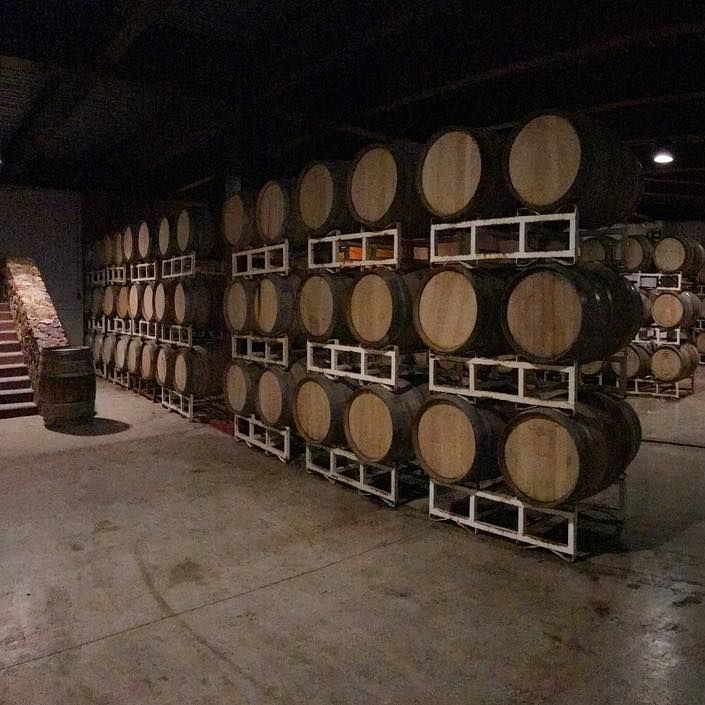 Bodega de vinos con poca luz, con barricas de roble apiladas sobre estantes metálicos y suelo de hormigón.