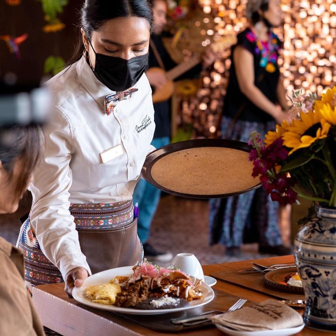 Un camarero enmascarado coloca un plato de comida en una mesa de un evento festivo con flores y luces cálidas.