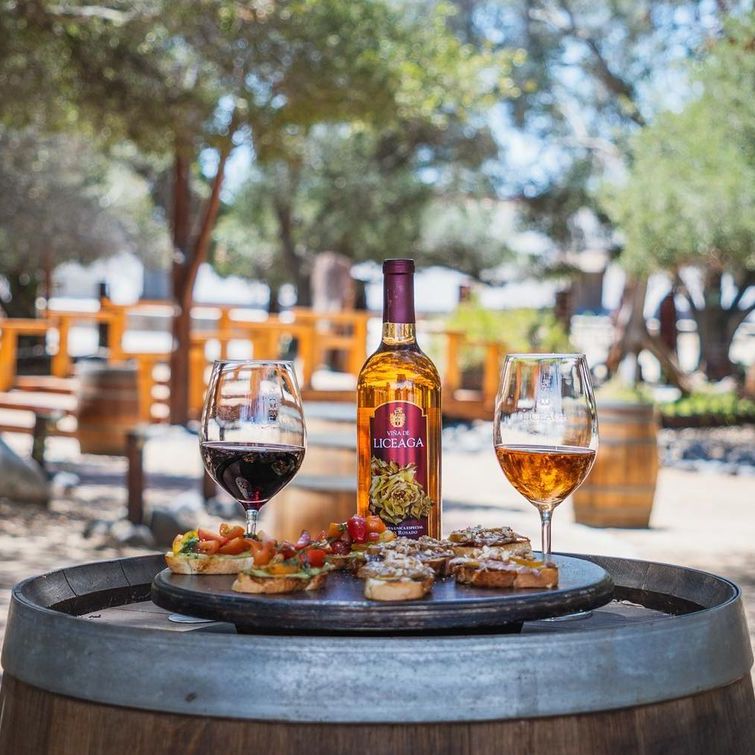 Vino y aperitivos sobre una mesa de barril al aire libre, con una botella y dos copas en primer plano.