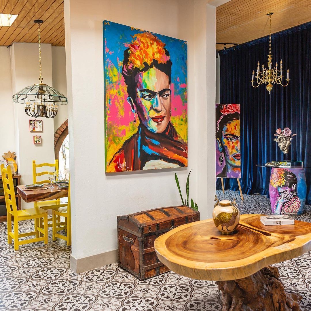 Habitación luminosa y llena de arte con un retrato colorido, suelo estampado, mesa redonda de madera y sillas amarillas.