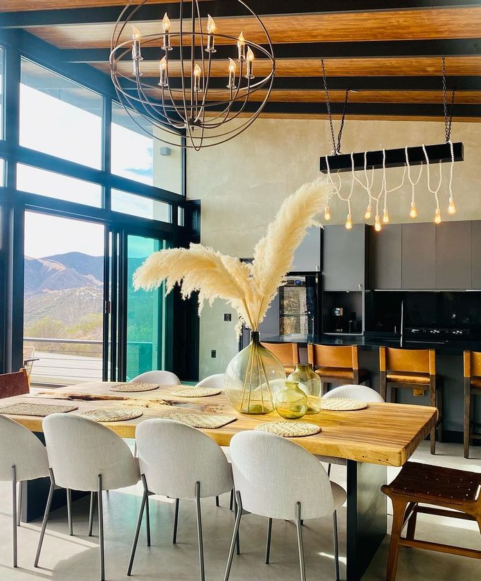 Comedor moderno con mesa de madera, sillas blancas, grandes ventanales y un jarrón con hierbas secas.