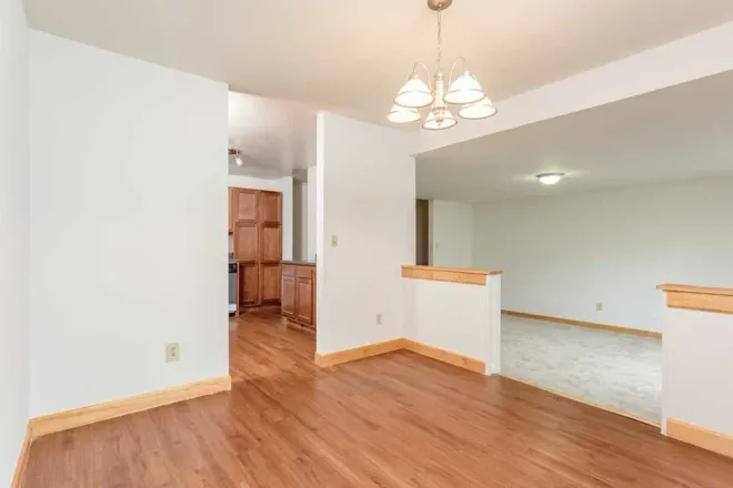 An empty interior space with light wood-look flooring, white walls, wood trim, and a chandelier over a dining area.
