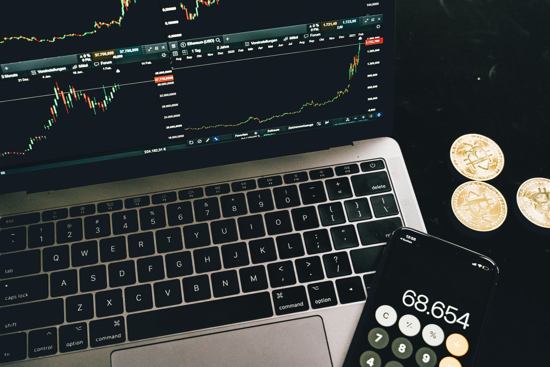 A laptop displaying financial charts next to a calculator showing 68,654 and three gold coins on a black surface.