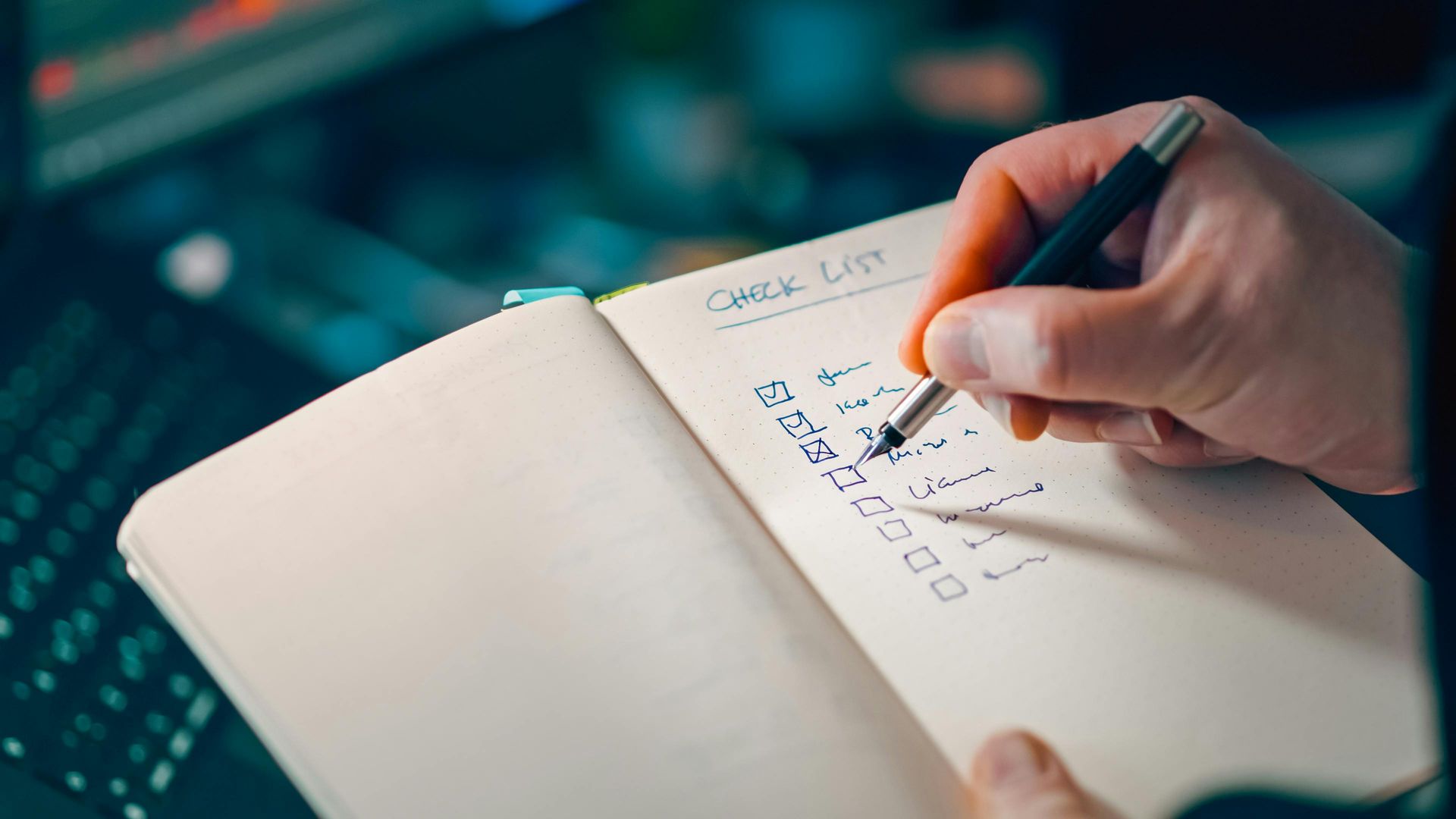 A person marks items off a handwritten checklist in a notebook while working at a desk.