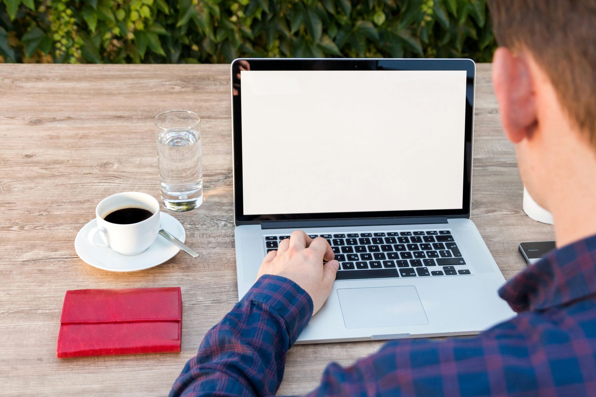 Person typing on a laptop at a wooden table with coffee, water, and a red notebook