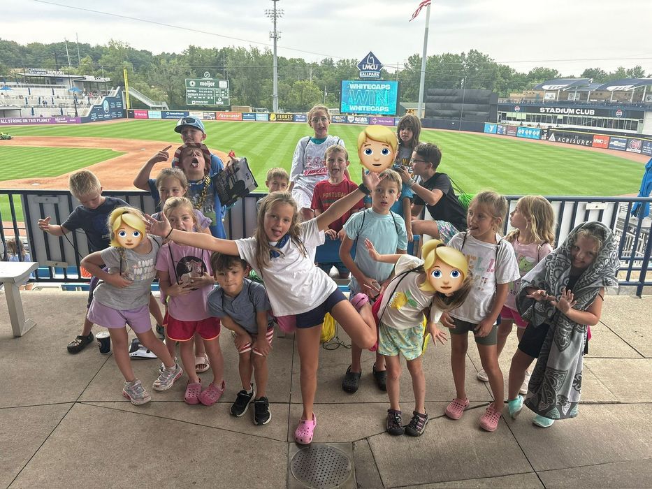 Children posing with mascot cutouts at a baseball stadium, with the field and scoreboard in the background