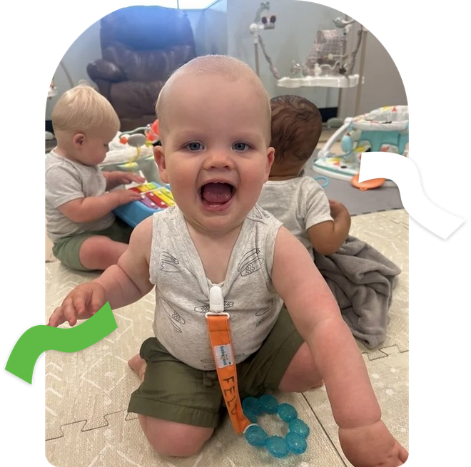 Happy baby sitting on a rug in a playroom, with toys and two other children in the background