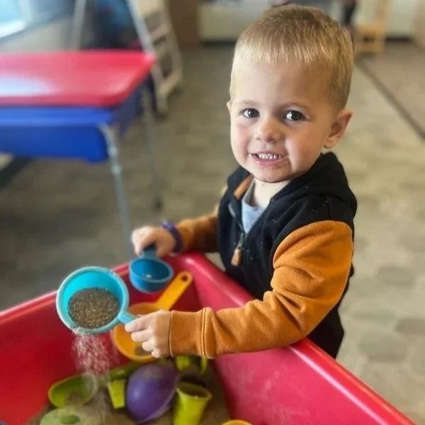Toddler smiling at a red sensory table, scooping with blue and yellow cups in a classroom.