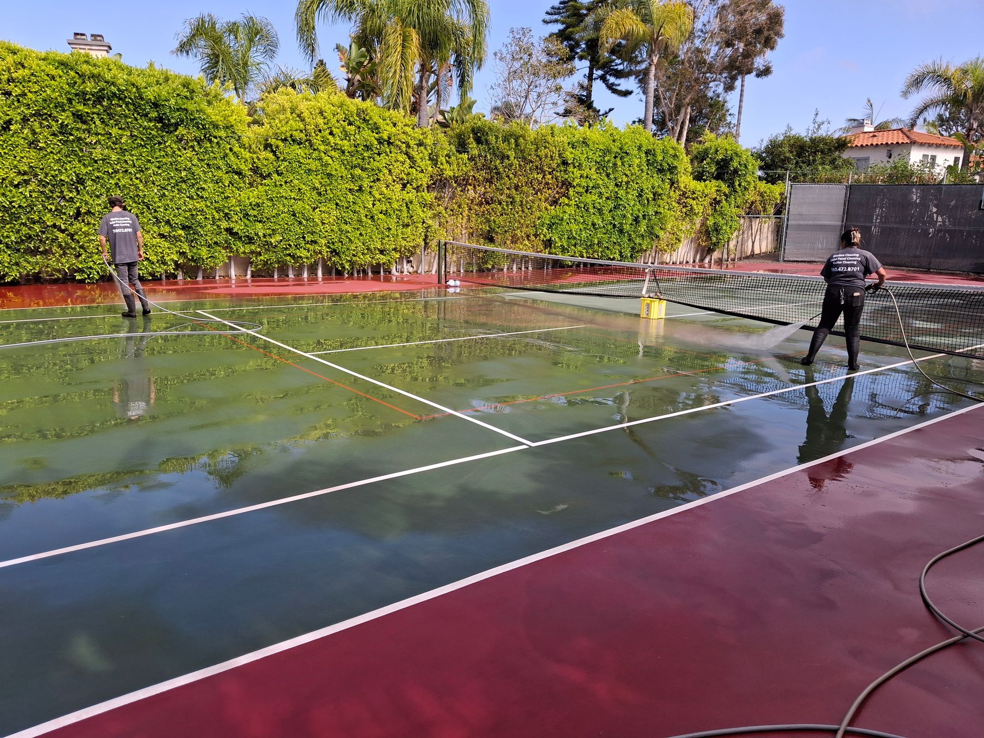 Two people power washing a wet, red and green tennis court.
