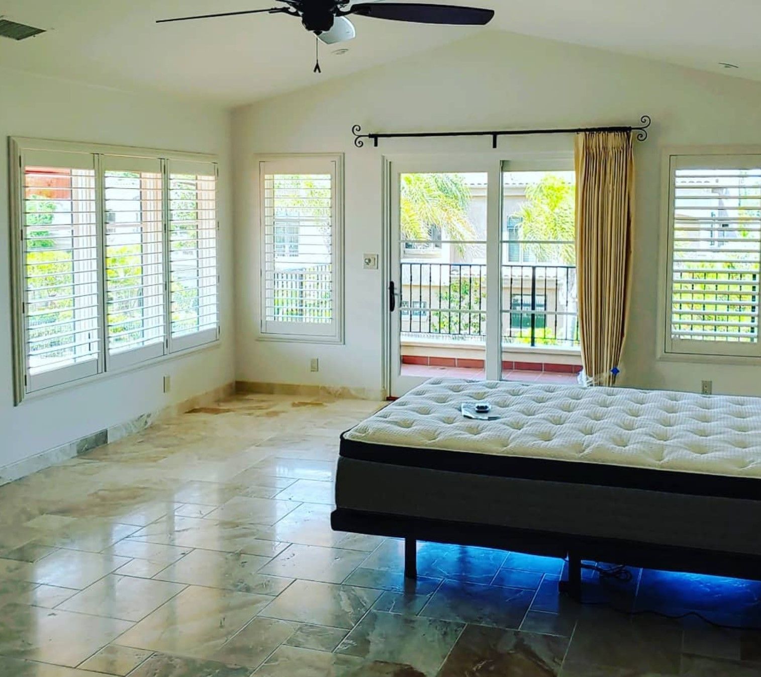Empty bedroom with shutters, a bed, and a balcony. Light brown tiled floor.