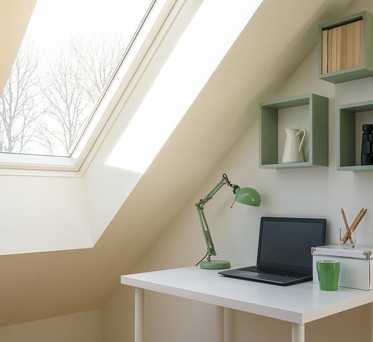 A laptop and green desk lamp sit on a white desk in a room with a skylight and green shelves.