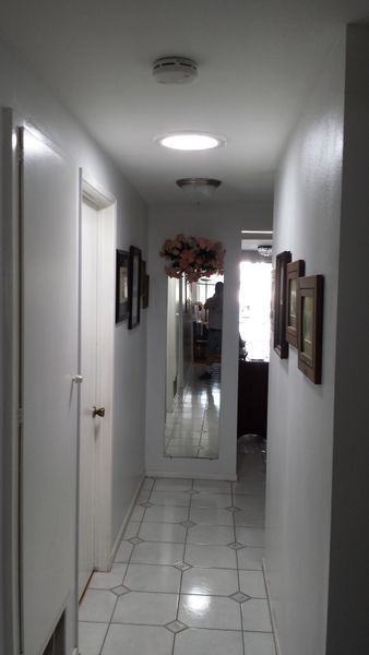 Narrow, white hallway with skylights, tiled floor, and framed art. A large mirror and closed door are visible.