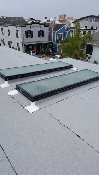 Two rectangular skylights on a gray flat roof, surrounded by white sealant. Buildings in background.
