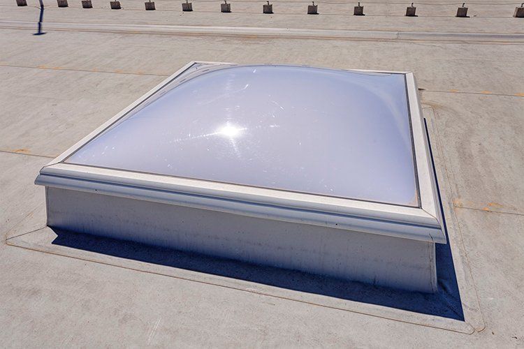 Skylight on a flat roof, white frame, clear dome, sunlight reflecting.