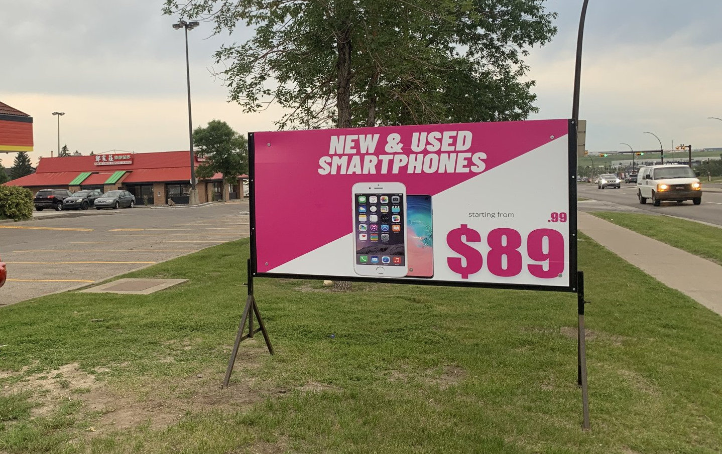 Sign advertising new and used smartphones for $89, set up on grass near a road and restaurant.