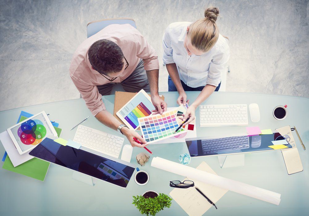Two people collaborate around a desk, examining color palettes and printouts, using computers.