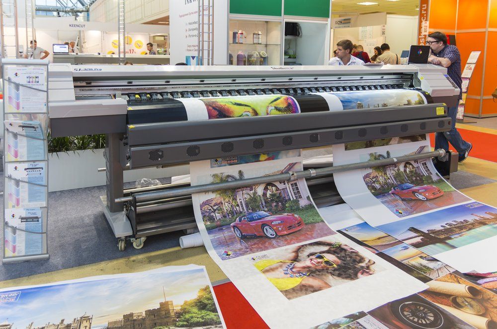 Large-format printer displaying colorful prints, including a car and portraits, in a trade show setting.