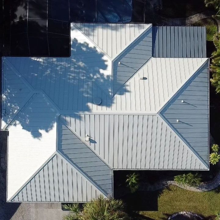 Aerial view of a house roof with light gray metal panels and tree shadows.