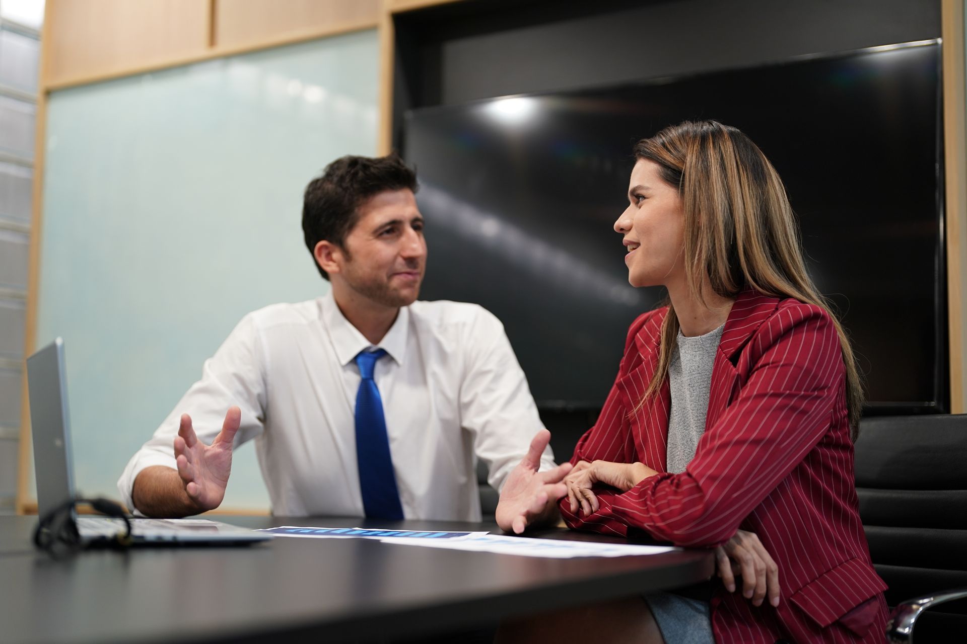 Two people talking at a conference table, with a white-shirted man and red-blazer woman in an office
