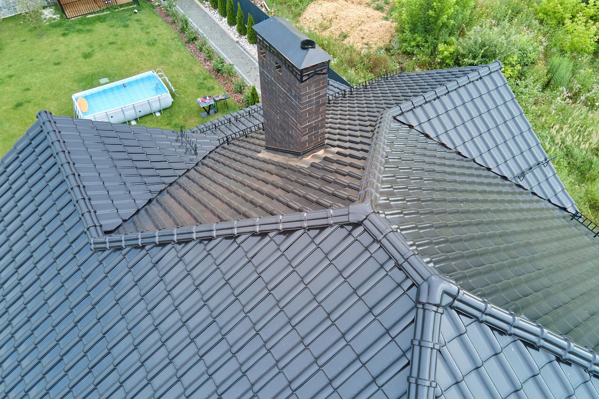 Aerial view of a dark gray shingled roof with a brick chimney and green yard below