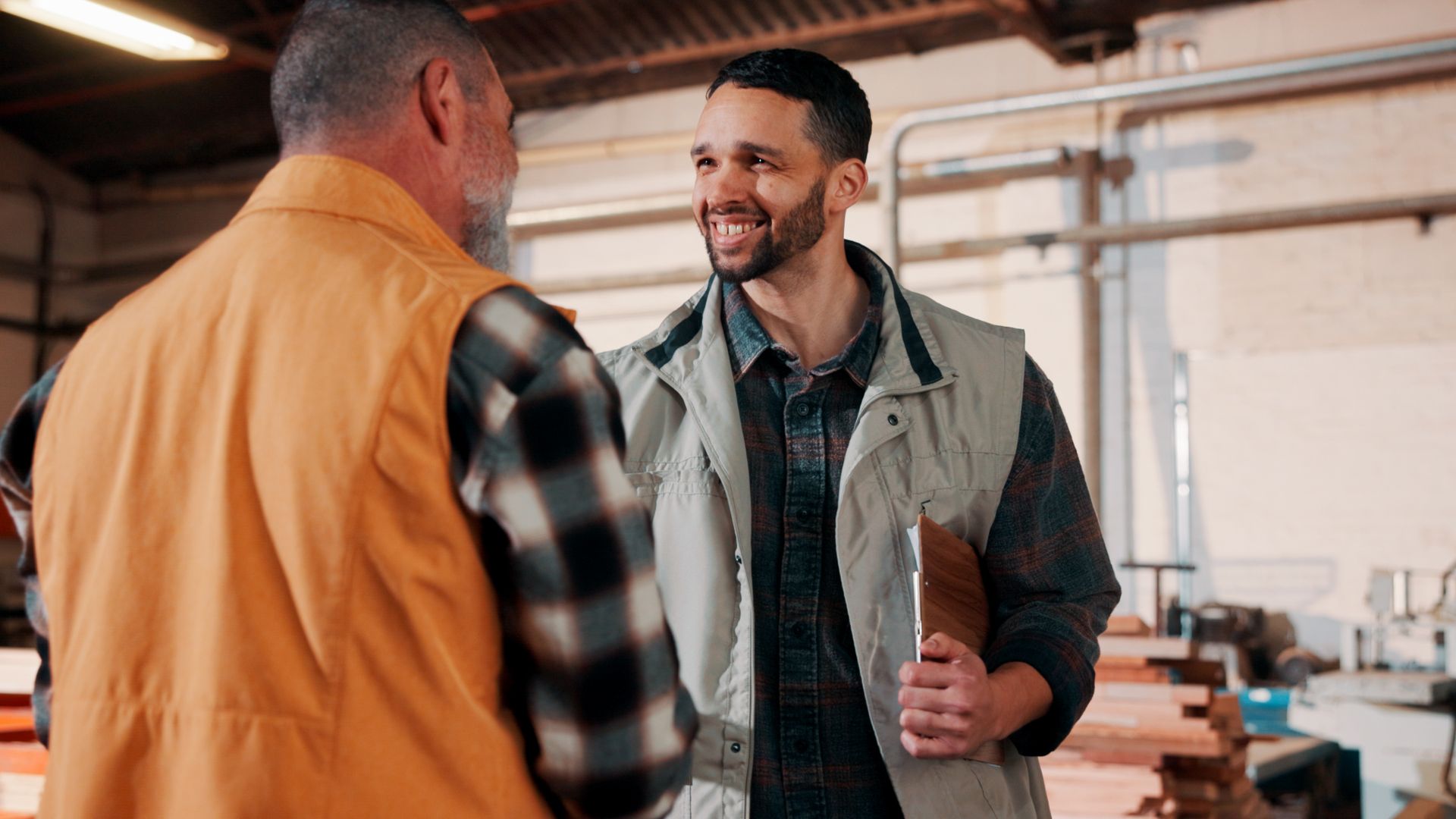 Two workers talking in a workshop, one wearing an orange vest and the other holding a tablet.
