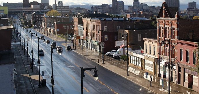 View of a city street with wet pavement, lined with buildings and streetlights, with a distant cityscape.