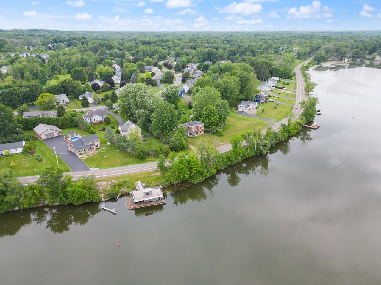 Aerial view of houses and a road along the edge of a lake, with a treeline and cloudy sky.