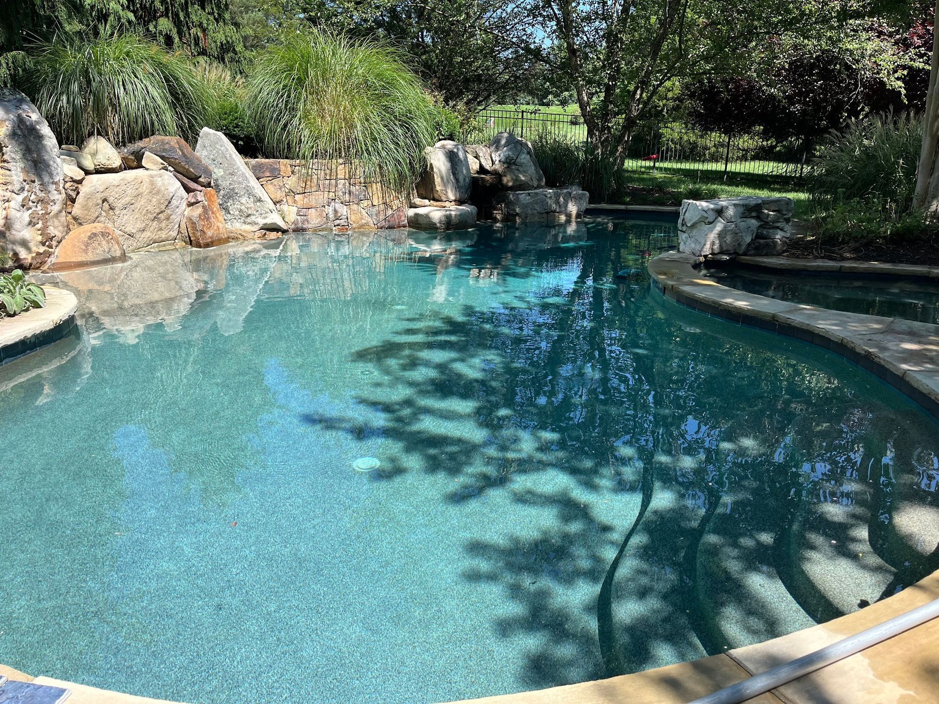 Swimming pool with rock waterfall feature, steps, and surrounding greenery.