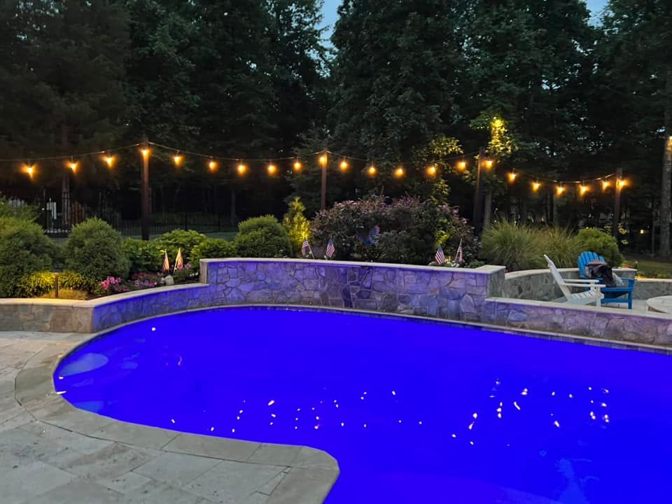 Nighttime pool scene with string lights, blue water, and stone wall.