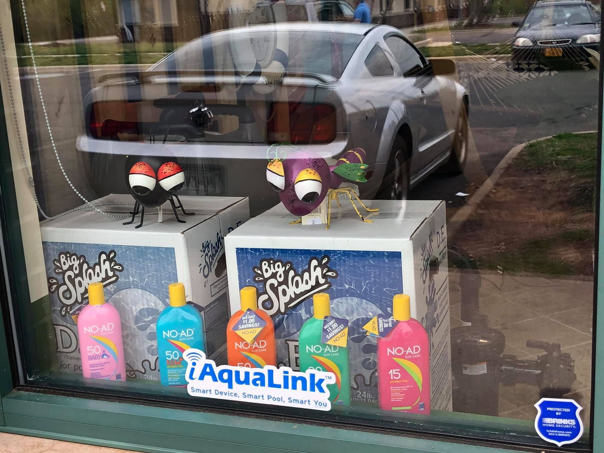Store window display with toy creatures, cleaning products, and silver car reflection.