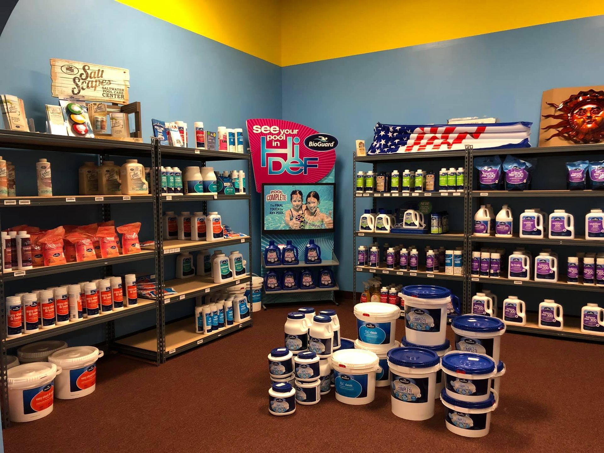 Pool supply store interior with shelves stocked with chemicals, supplies, and displays. Red, white, blue color scheme.