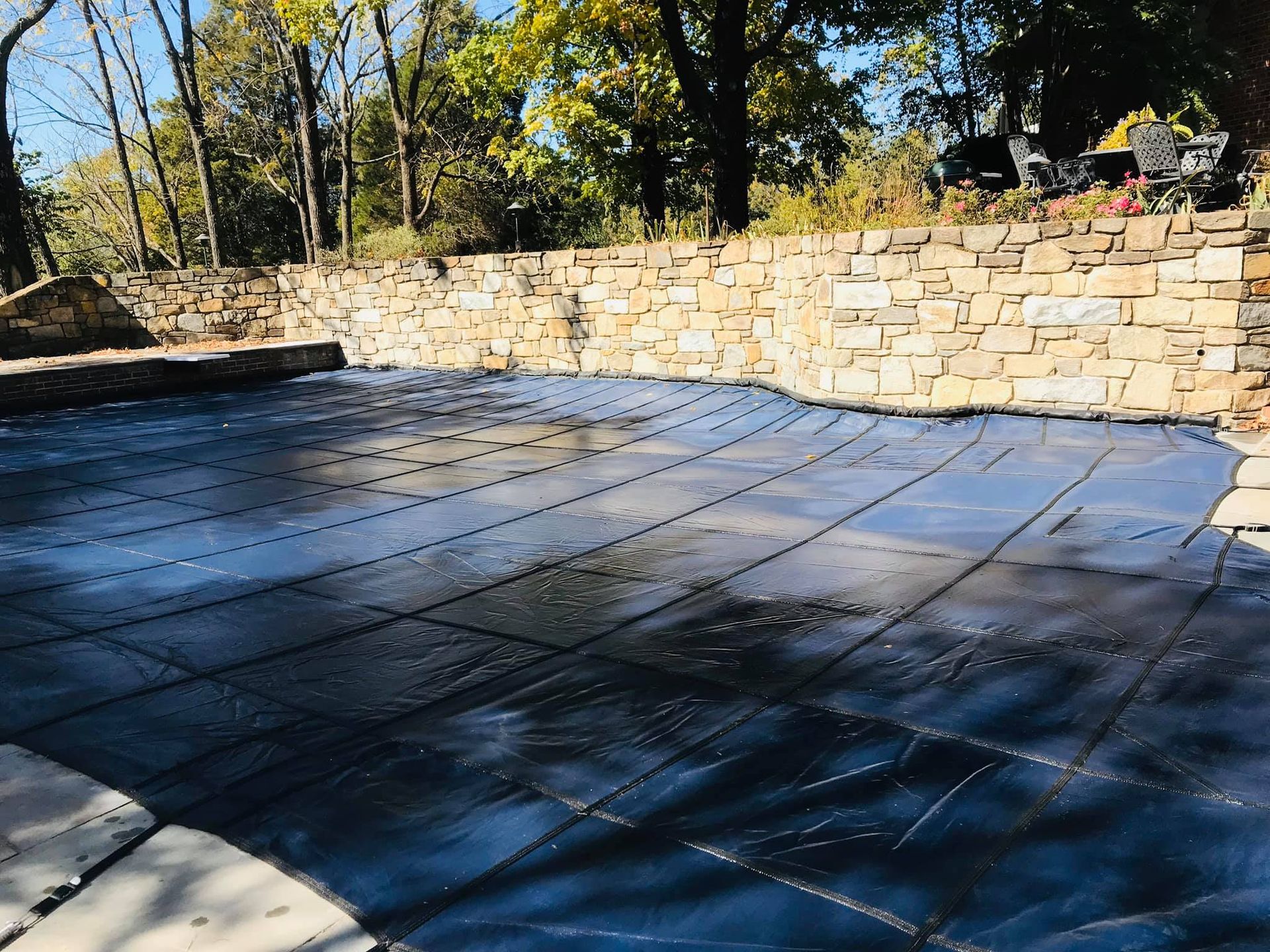 Pool covered with a dark tarp; stone wall in the background, trees in the distance.