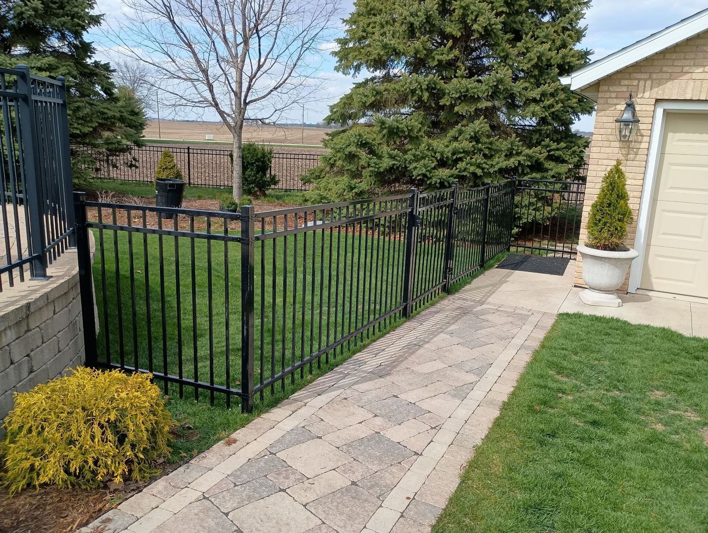 A brick walkway leading to a house with a black fence.