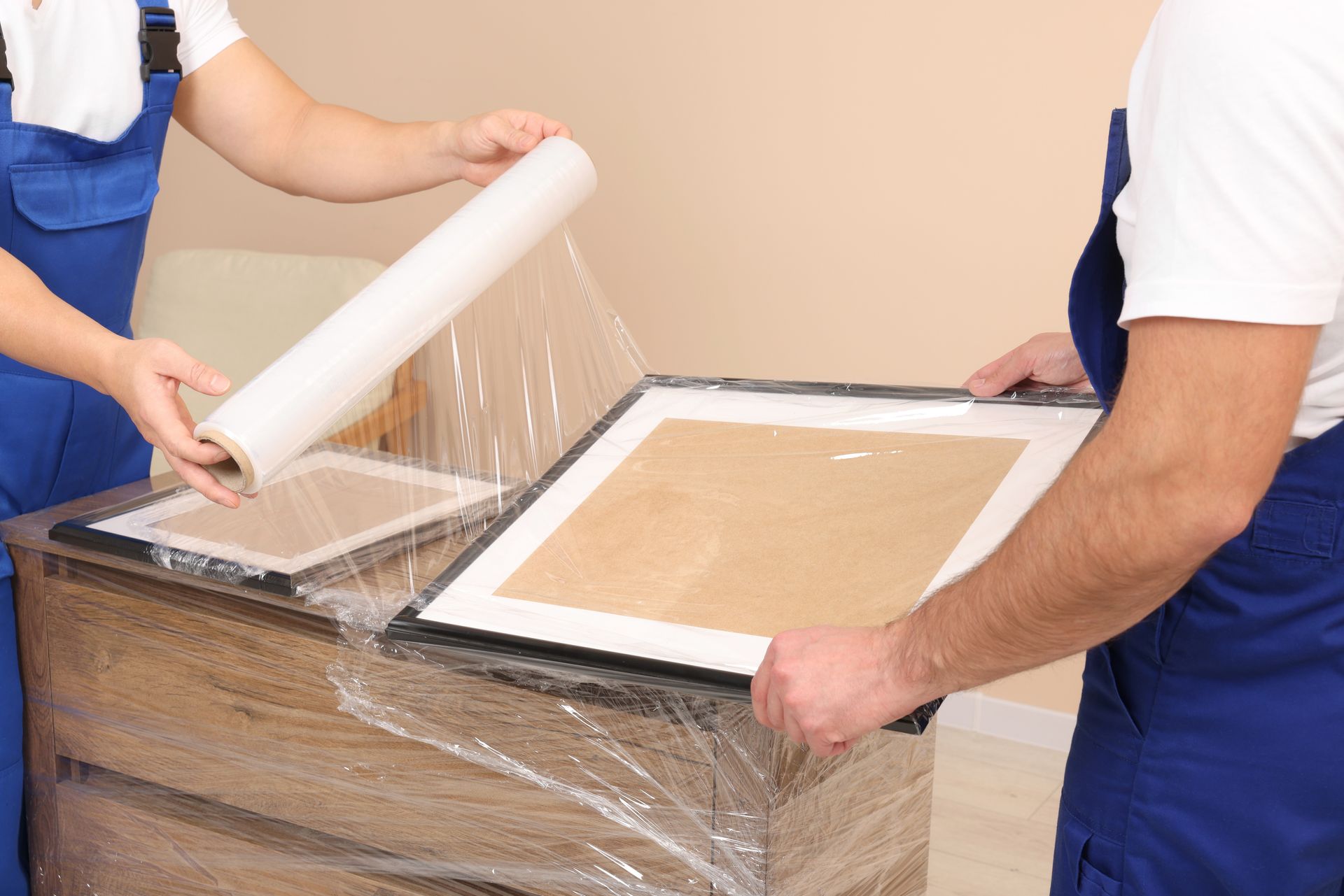 Two movers wrapping framed artwork in clear plastic.
