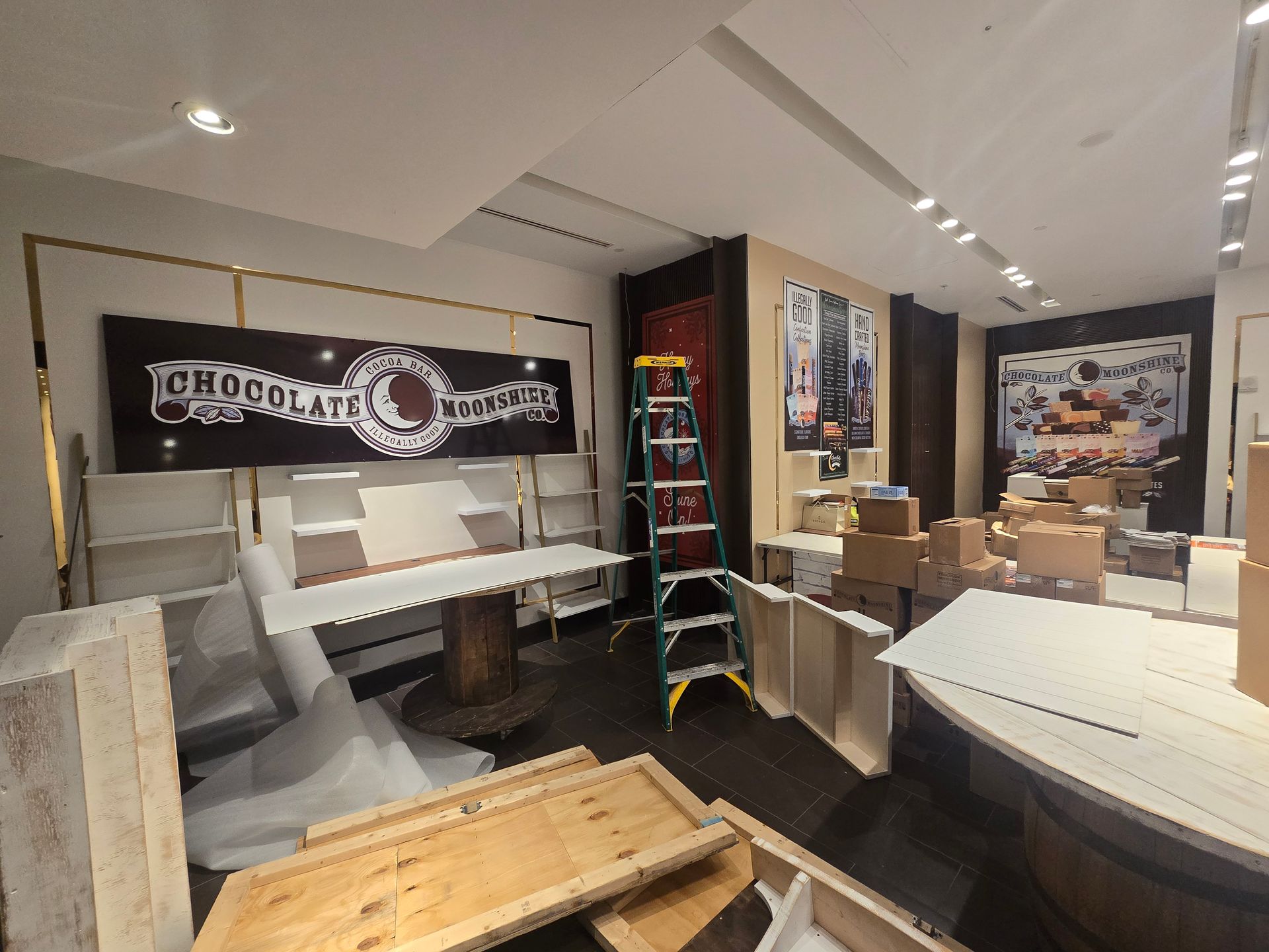 Indoor retail display with white counters, stacked boxes, banners, and a small ladder in a showroom-like space