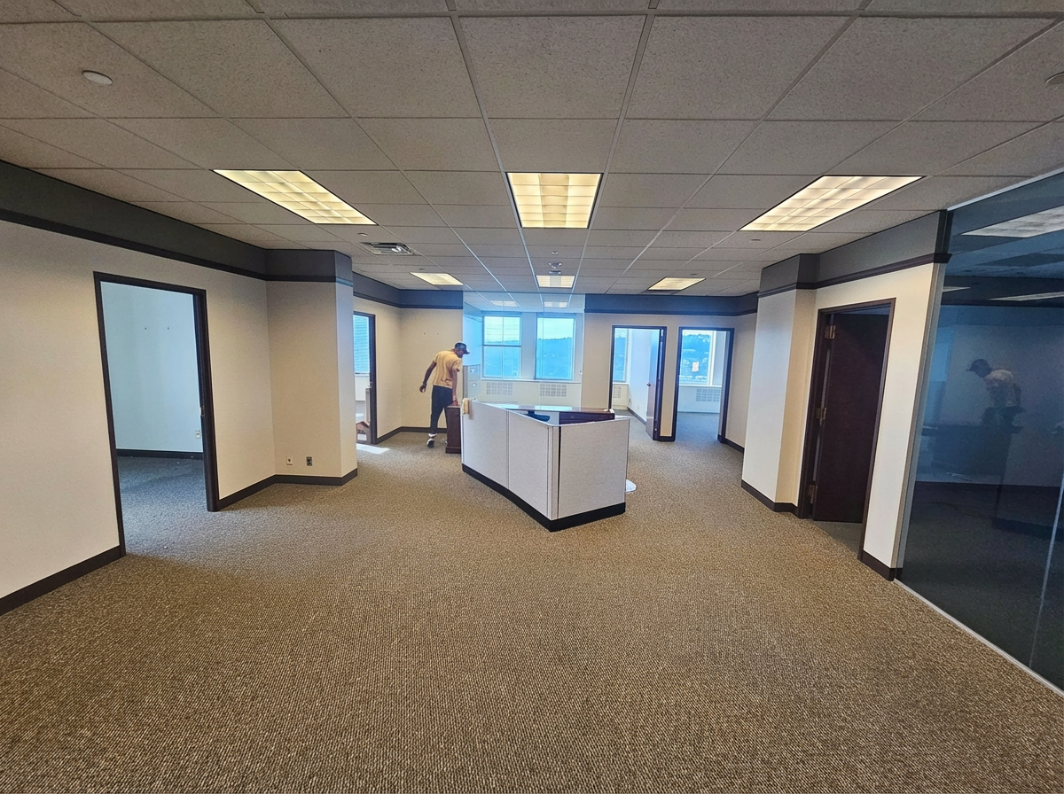 Empty office space with brown carpet, multiple doorways, and a person walking towards a window.