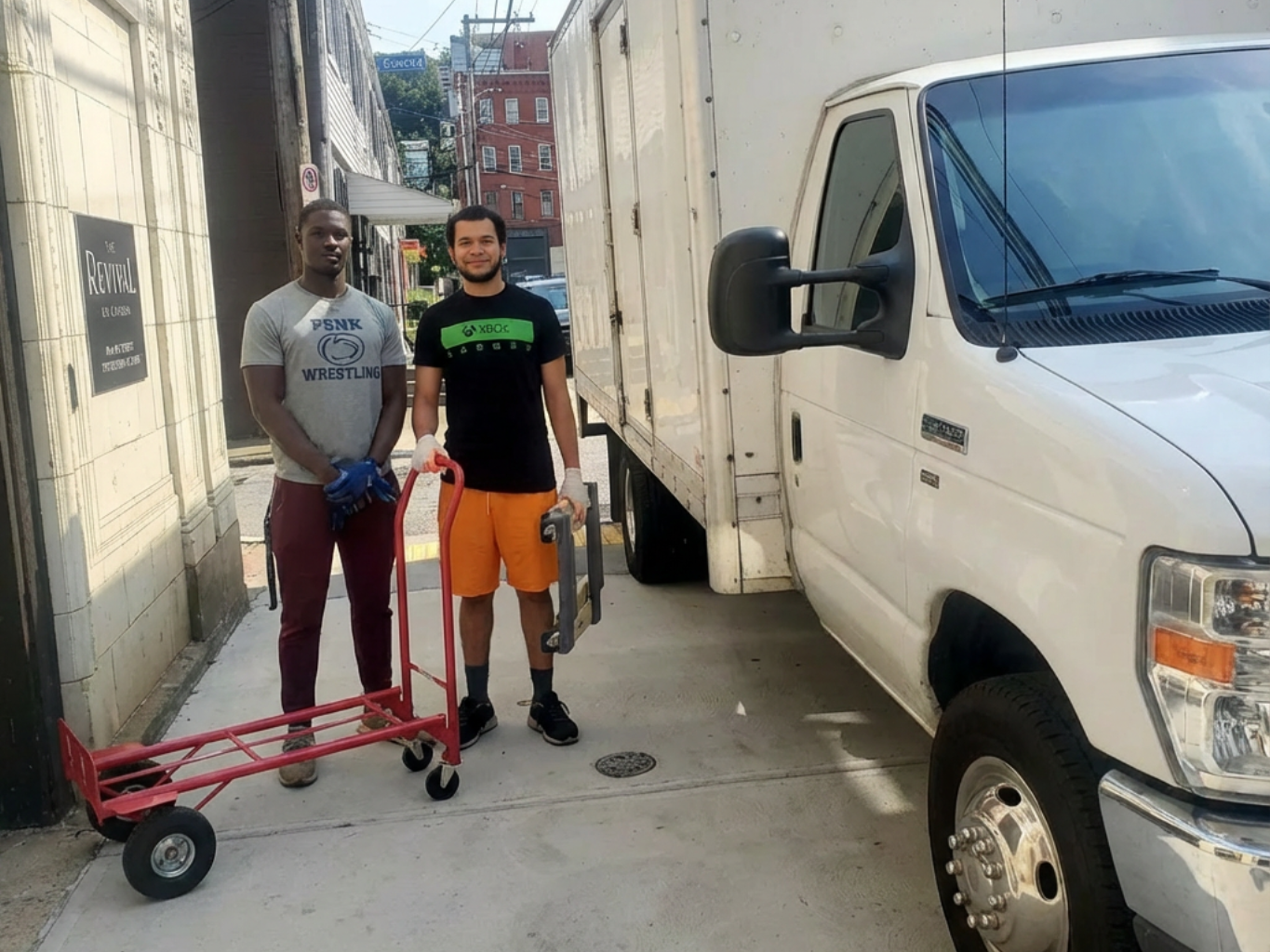 Two people stand beside a white box truck in a narrow alley, next to a red hand truck.