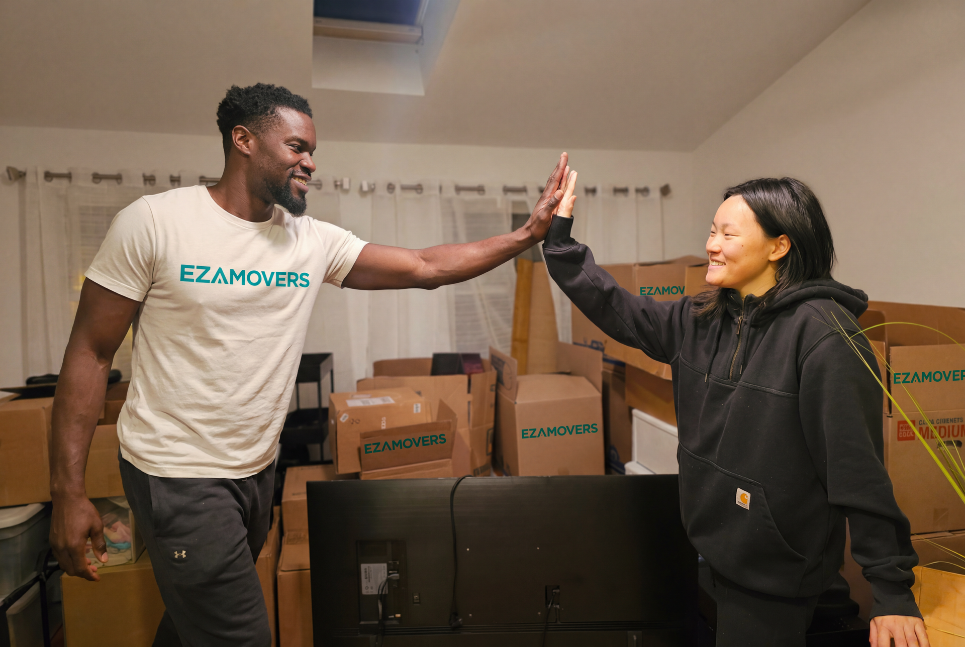 Man and woman high-five in a room with moving boxes, likely after a successful move. The man wears a
