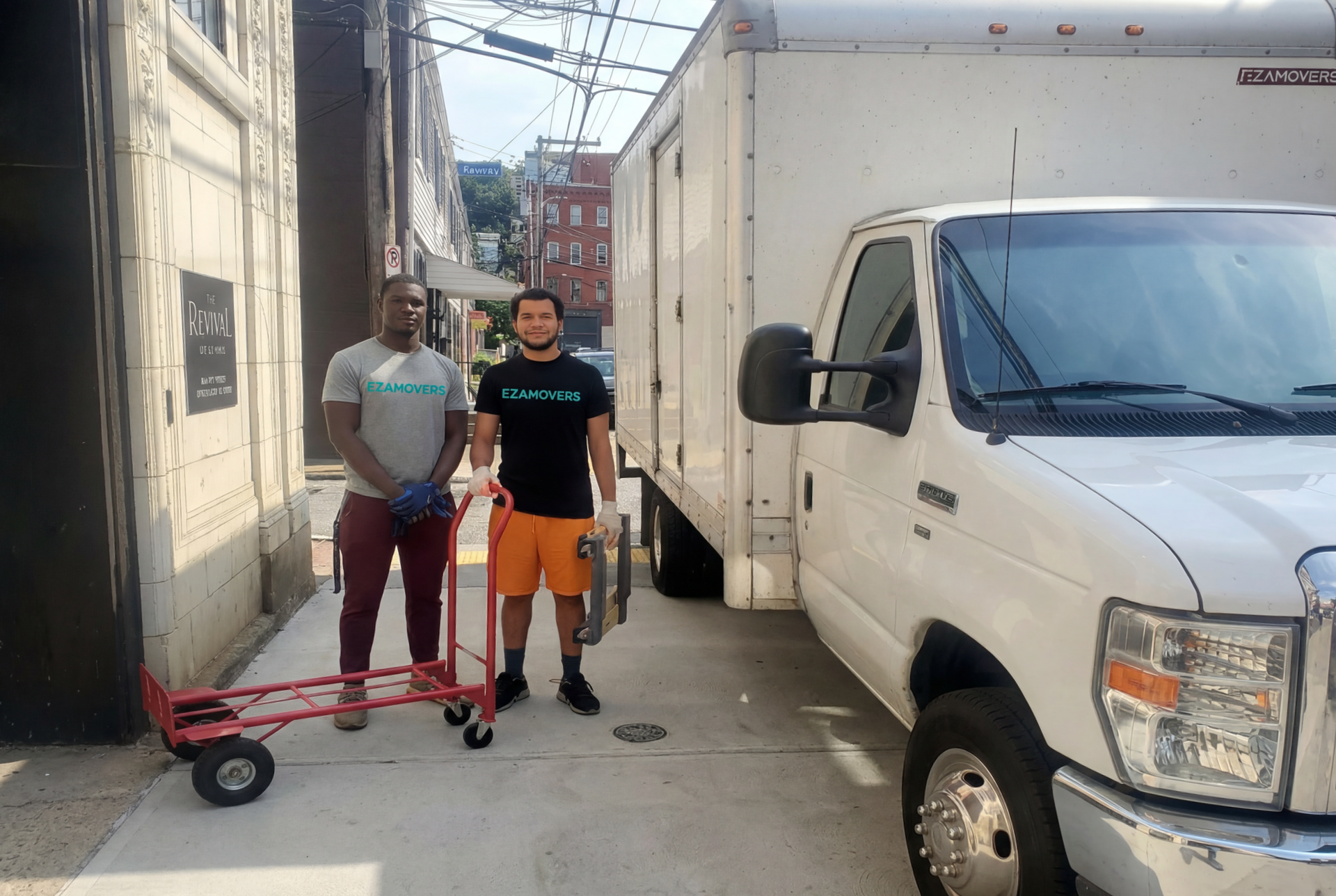 Two men standing beside a moving truck in an alley, one with a hand truck.