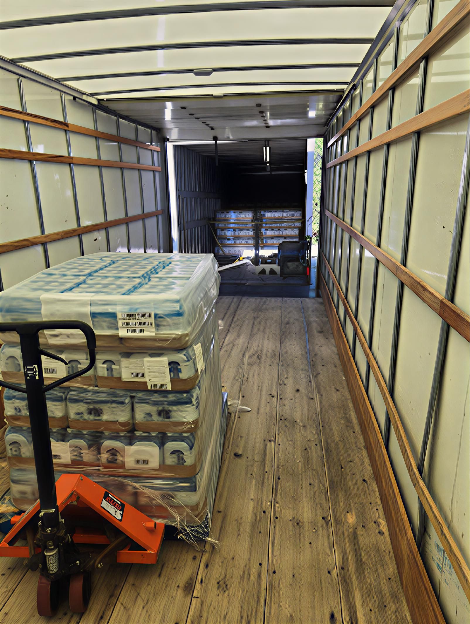 Inside a cargo trailer, a pallet jack holds a wrapped pallet of bottled water; the open back door shows additional water pallets.