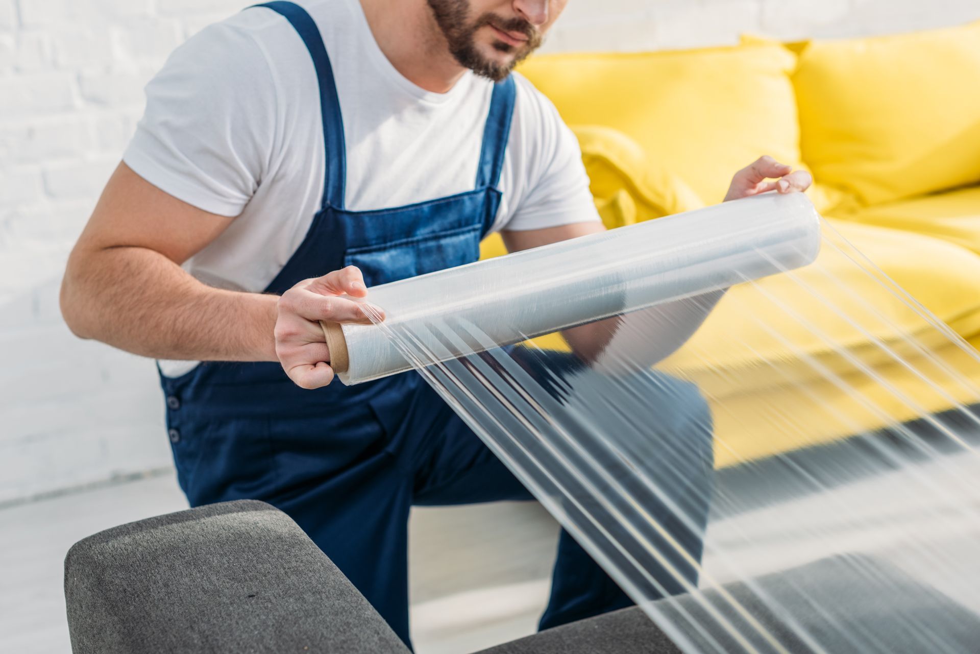 Man in blue overalls wrapping a gray chair with clear plastic wrap in a bright room.