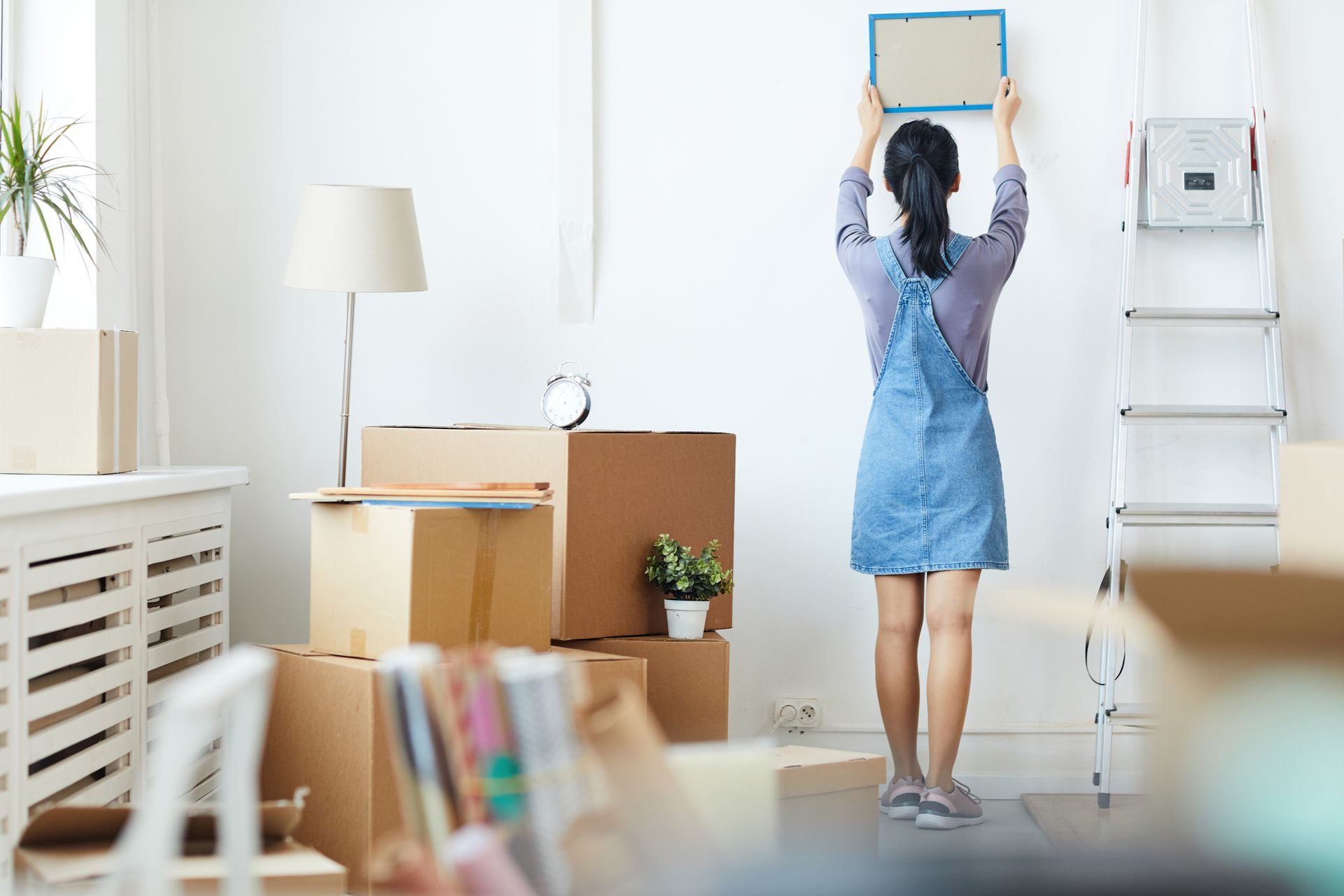 Woman hangs a framed picture on a white wall, standing on a ladder, surrounded by moving boxes in a bright room.