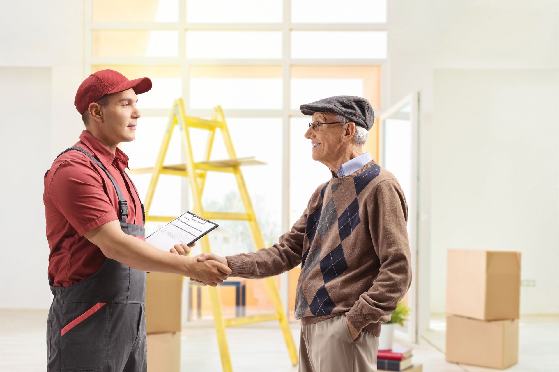 A mover in red overalls shakes hands with an elderly man, boxes and ladder in a bright room.