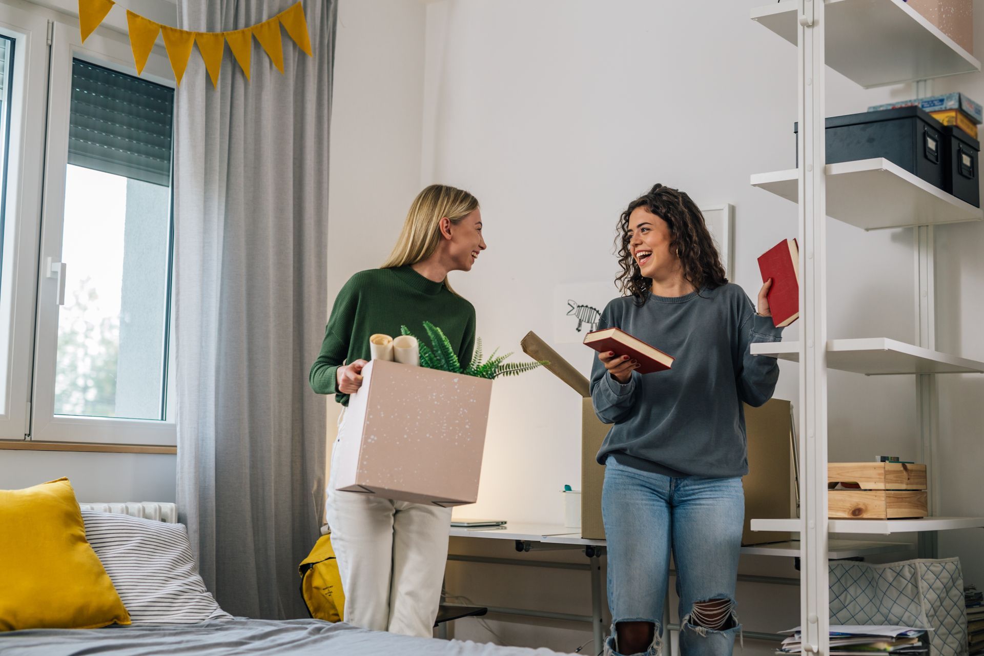 Two young women unpacking boxes in a brightly lit bedroom. One holds a box, the other a book; they are smiling and talking.