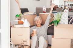 Smiling woman in a chair among moving boxes, holding a mug and raising her arm in a bright room