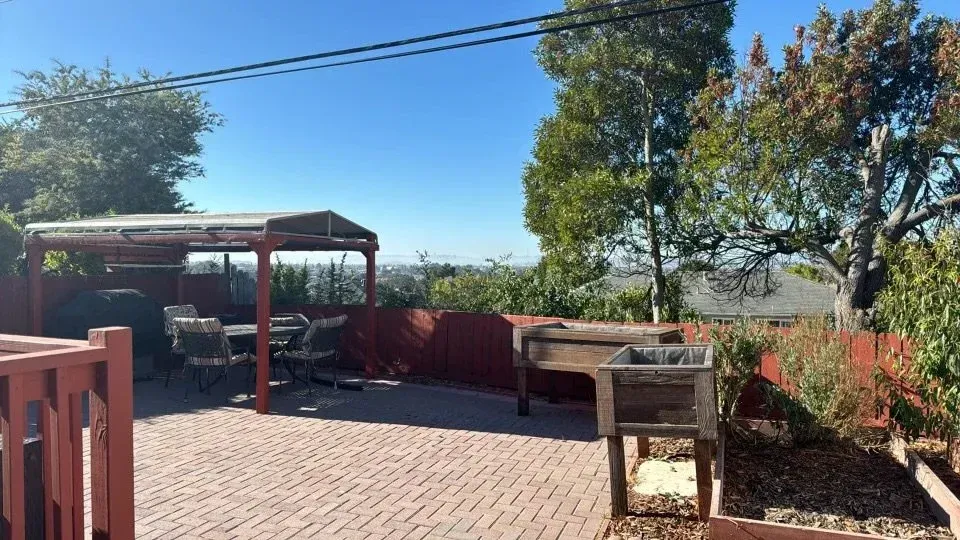Sunny backyard patio with brick paving, pergola, potted plants, and a barbecue grill near trees and shrubs