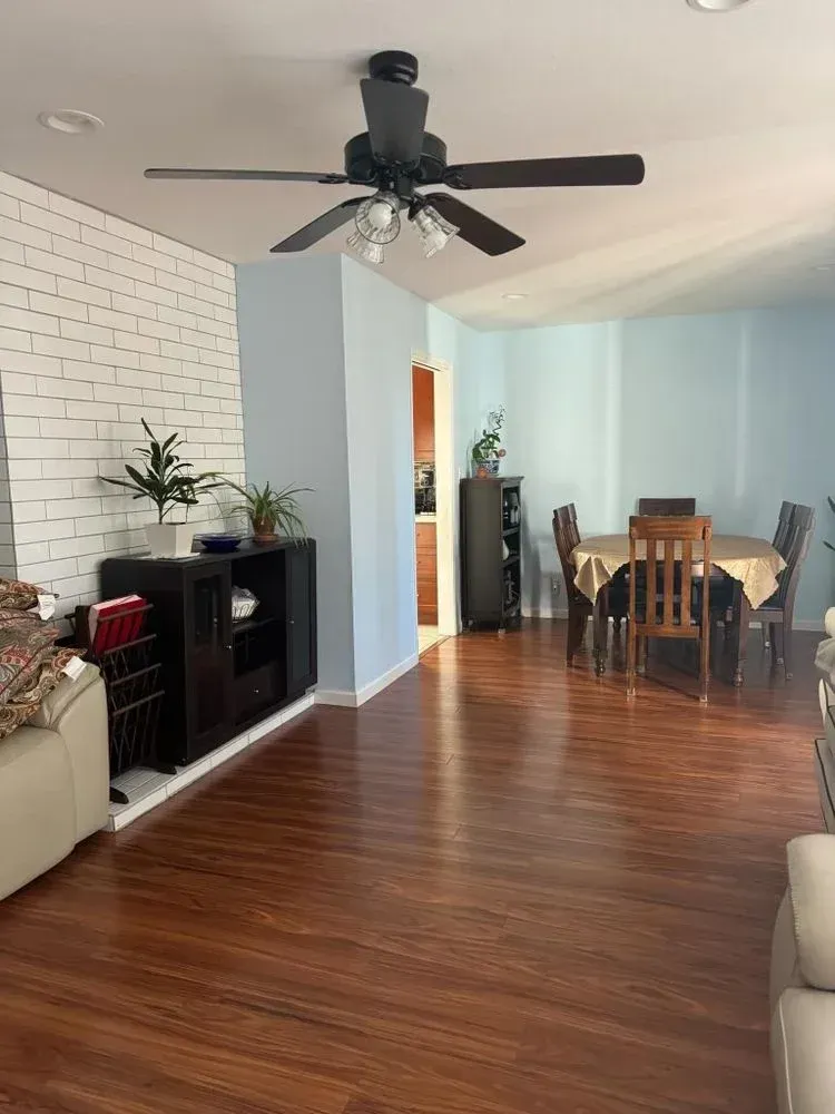 Living room with wood floors, black ceiling fan, white brick accent wall, and dining area in back