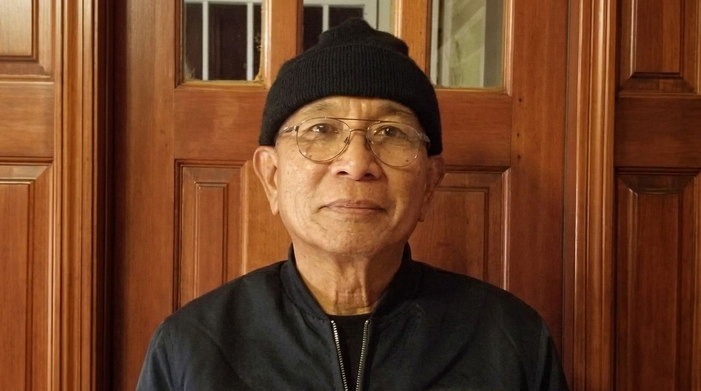 Person wearing glasses and a black beanie with a red-and-white logo, standing indoors against wooden doors.